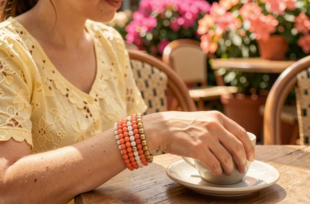 Bracelets en perles corail et dorées empilés sur le poignet d'une femme attablée à une terrasse de café en été